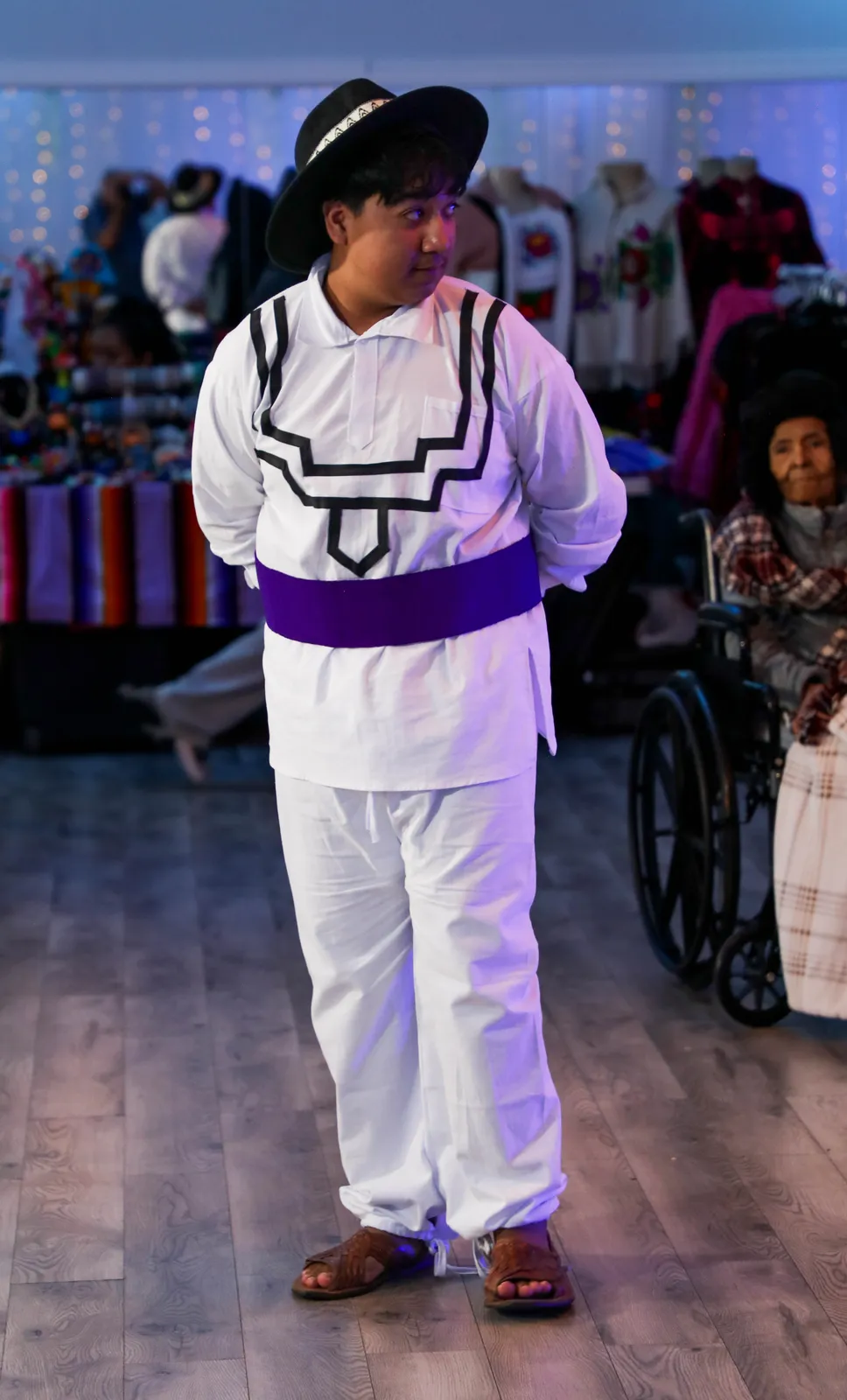 White clothing with dark geometric trim, a purple sash, and black hat at Oaxaca en Utah’s Fashion Show 2025