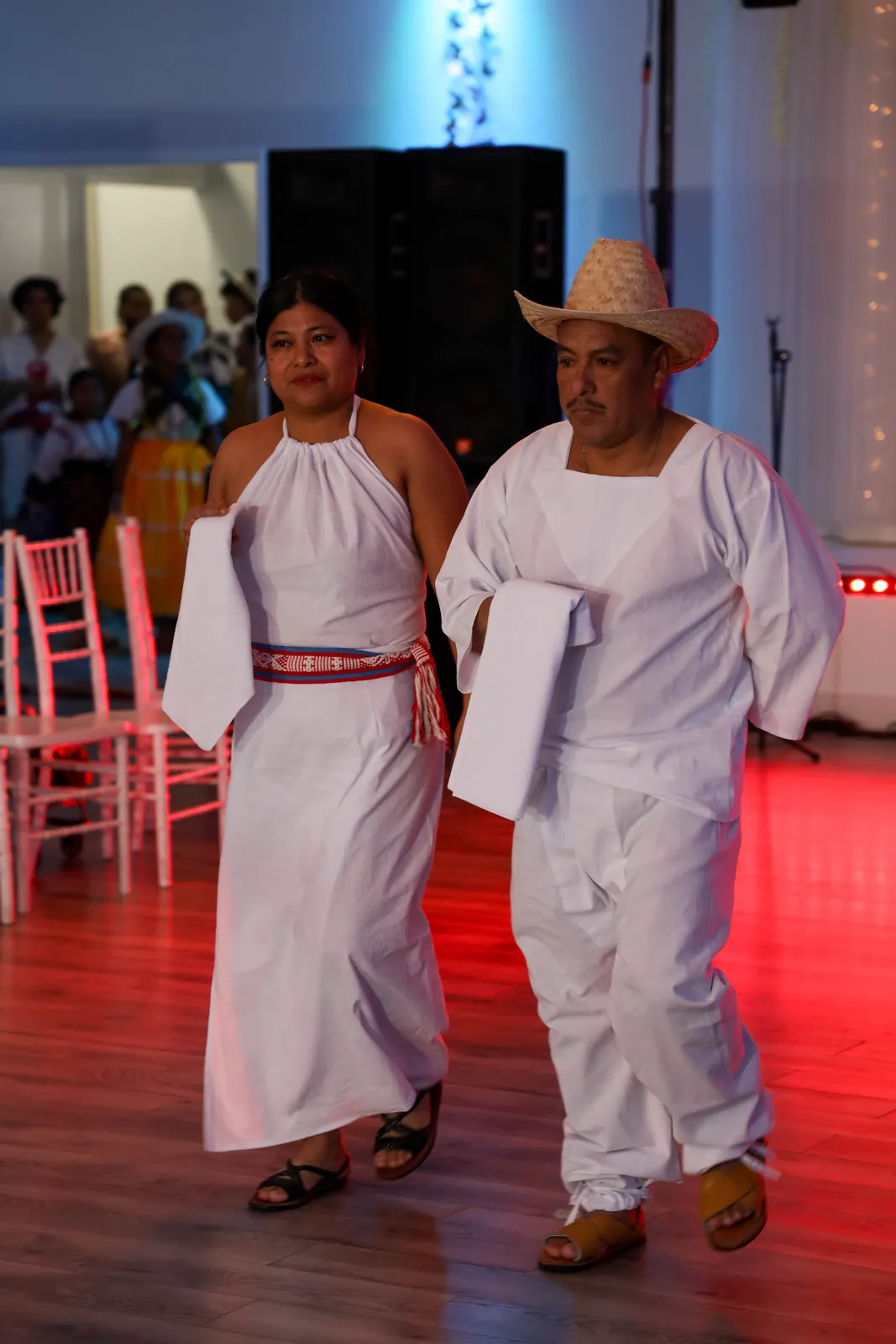 A pair presents white clothing, a woven red sash, and a palm hat at Oaxaca en Utah’s Fashion Show 2025