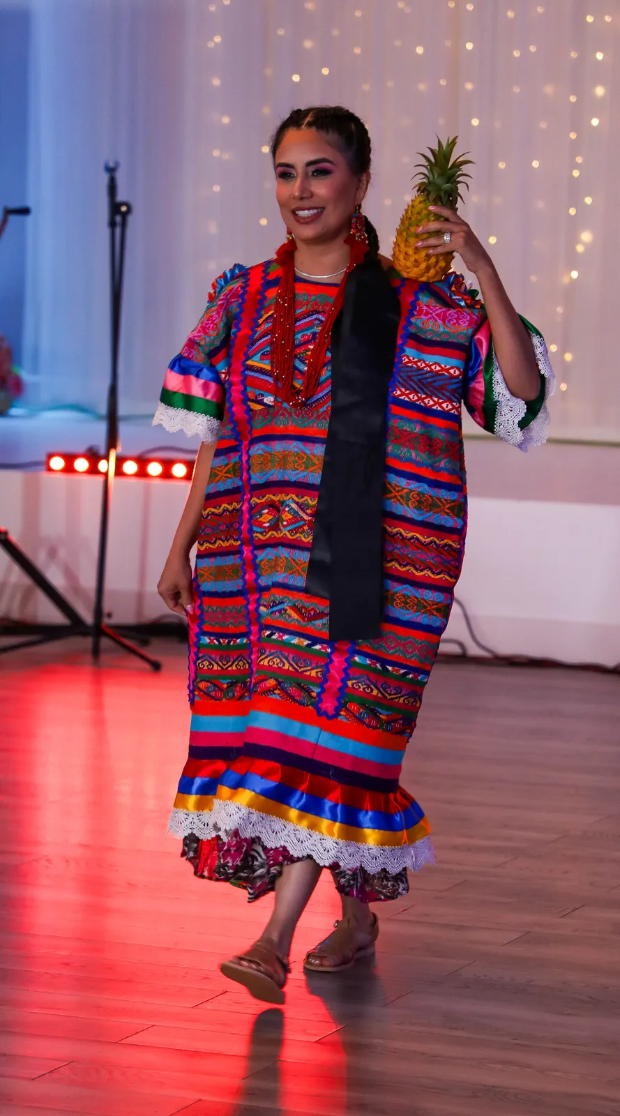 Multicolored geometric huipil with vertical panels, ribboned braids, and a pineapple at Oaxaca en Utah’s Fashion Show 2025