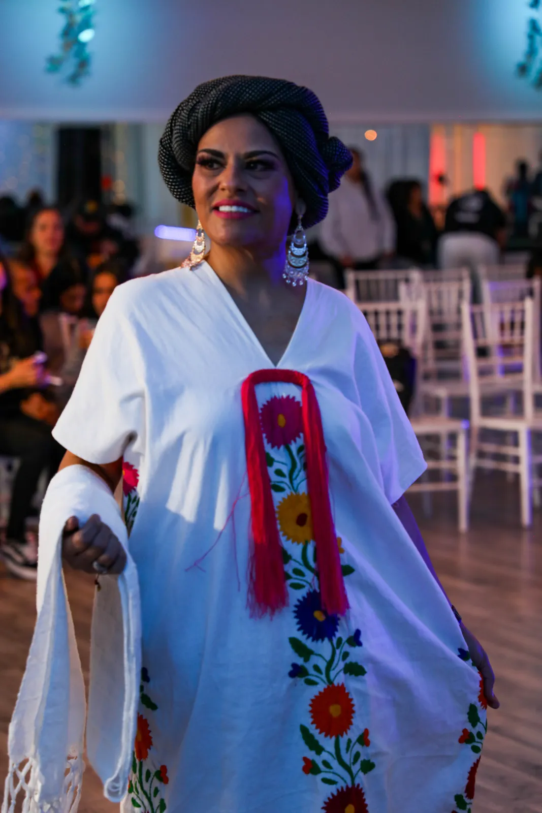 White huipil with a colorful vertical embroidered panel, wrapped skirt, and black wool headdress at Oaxaca en Utah’s Fashion Show 2025
