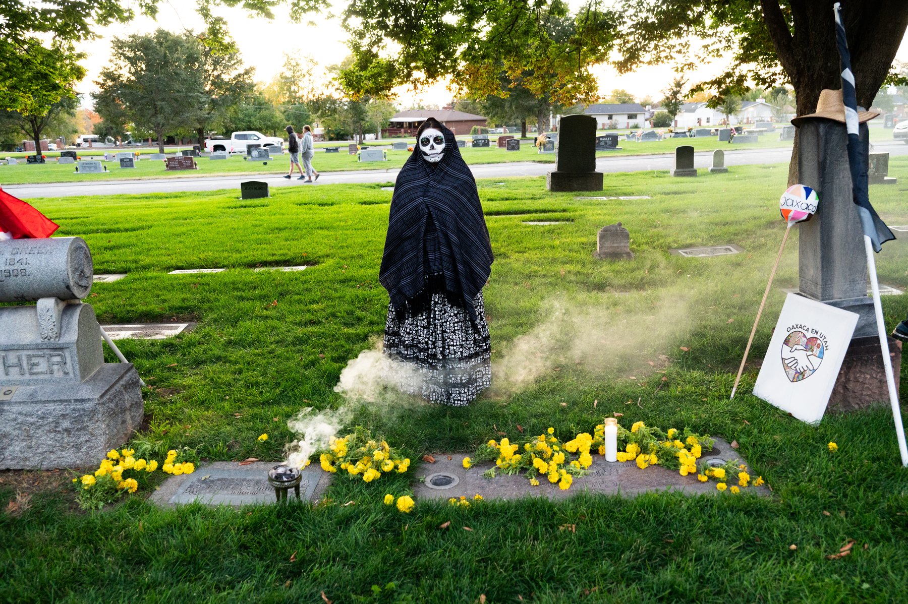 Una participante con chal negro y maquillaje de calavera está junto a una tumba con cempasúchiles y humo de incienso