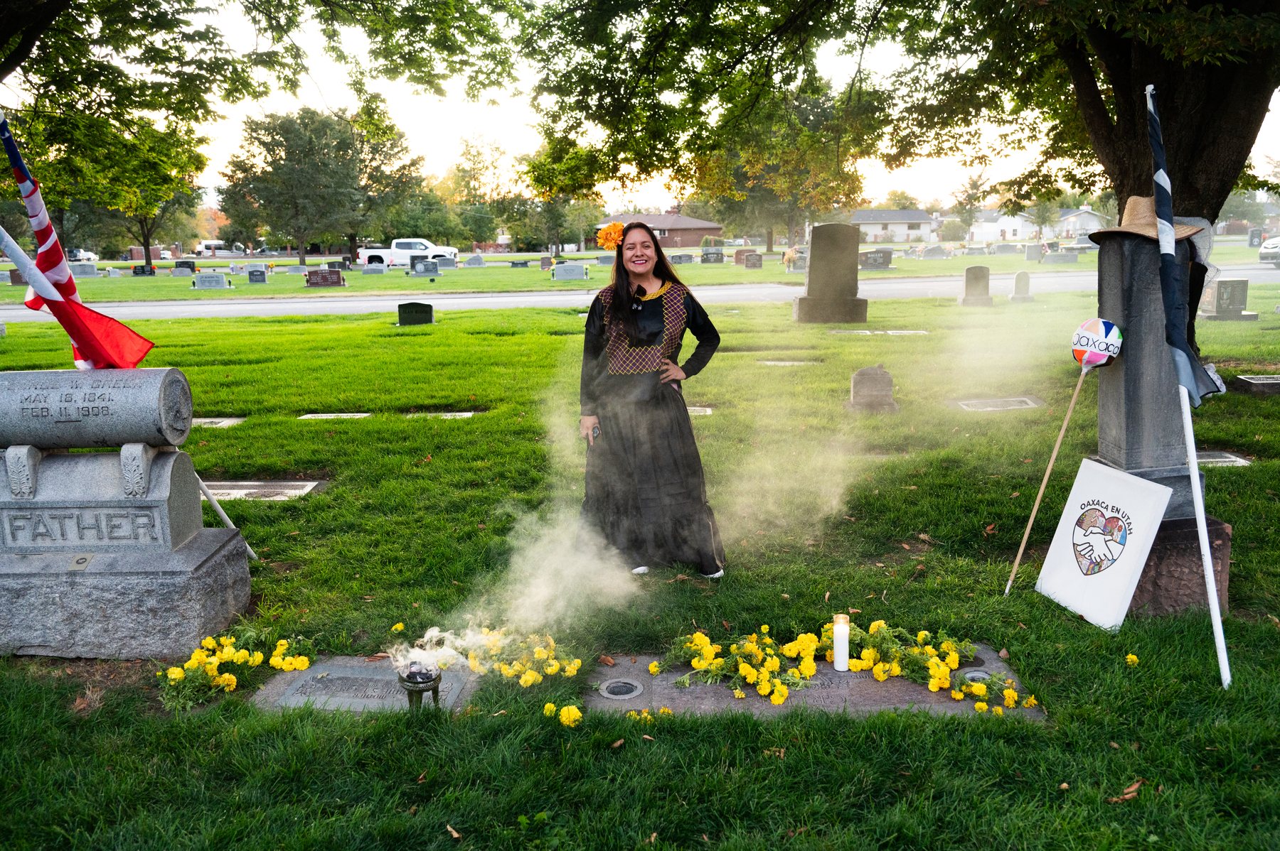 Una participante con vestido negro está junto a una tumba bordeada de cempasúchiles entre humo