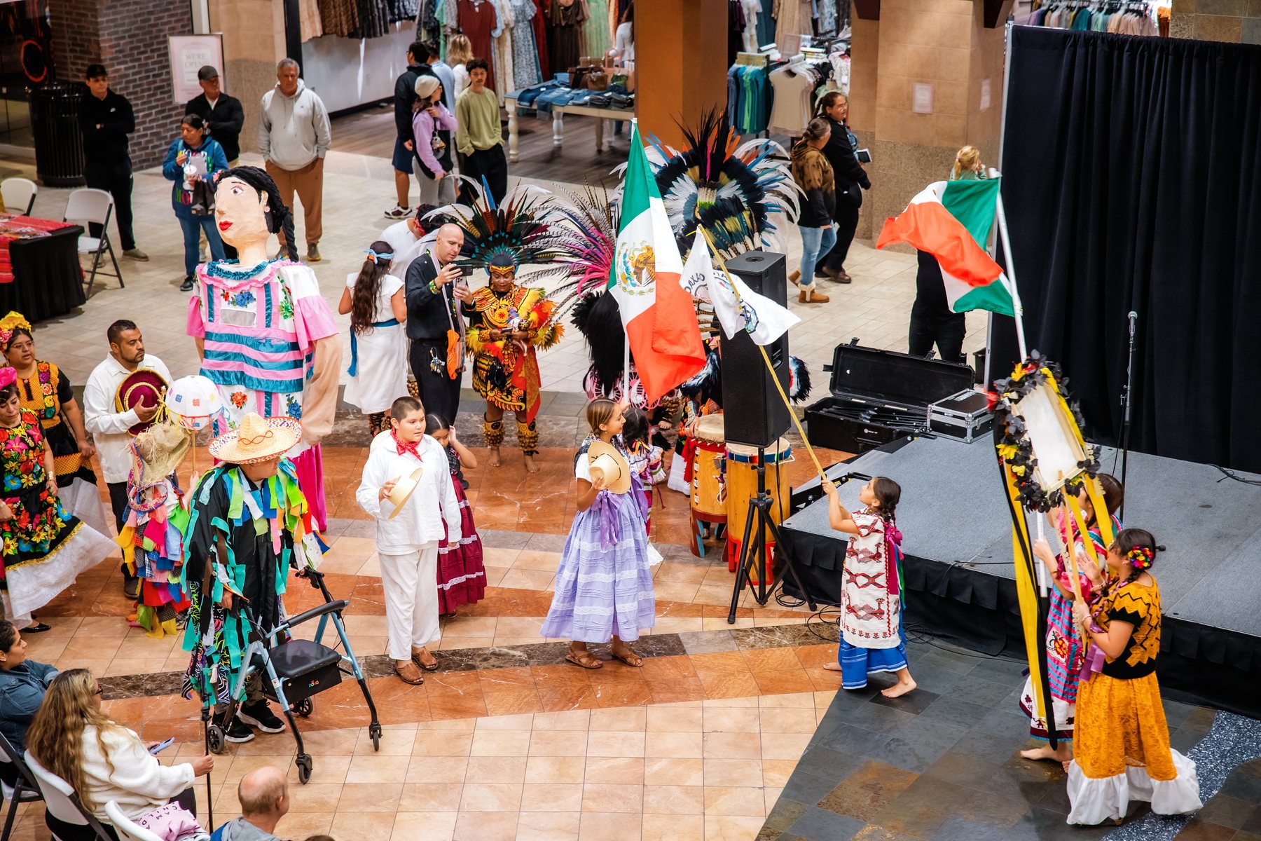Participantes con banderas, tocados de plumas y vestimenta colorida rodean el escenario del centro comercial