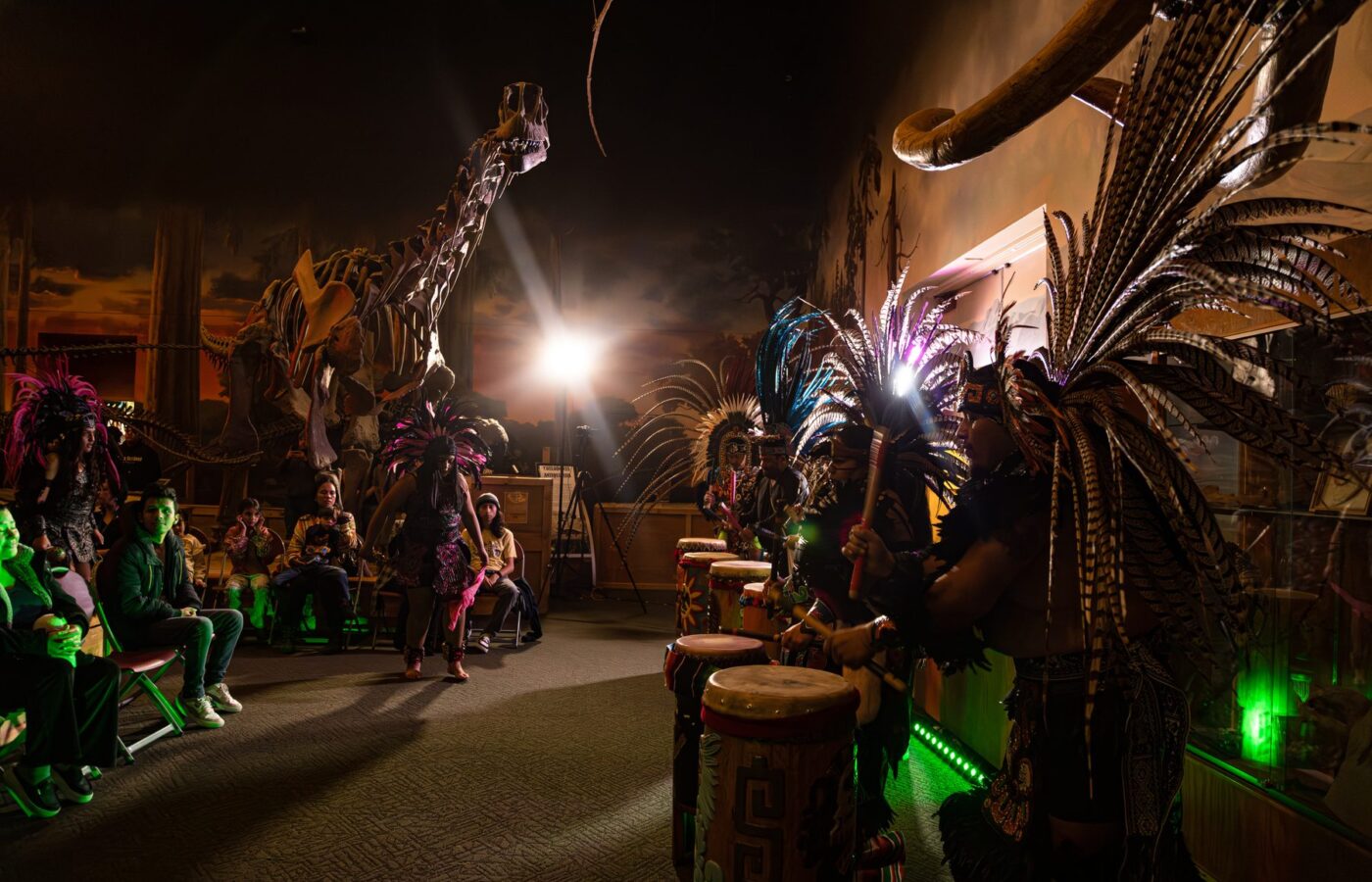 Integrantes de Danza Azteca bailan y tocan tambores bajo grandes esqueletos de dinosaurio mientras las familias observan