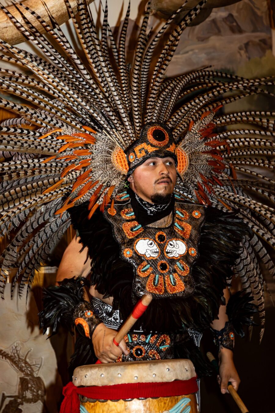 Un integrante de Danza Azteca sostiene una baqueta mientras porta un penacho de plumas negras, naranjas y blancas