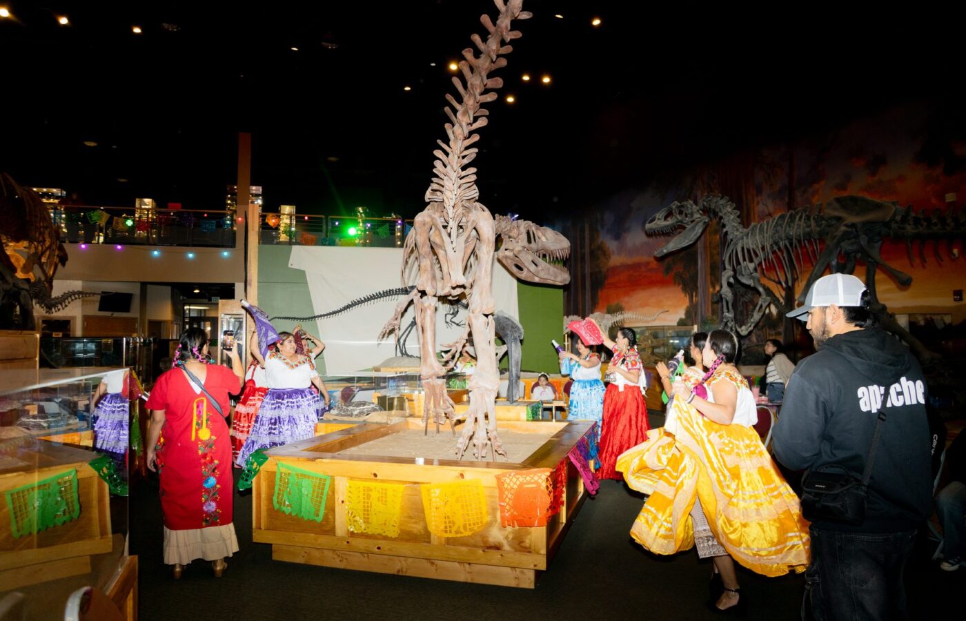 Mujeres con faldas coloridas bailan alrededor de un esqueleto de dinosaurio dentro de Ogden Dinosaur Park