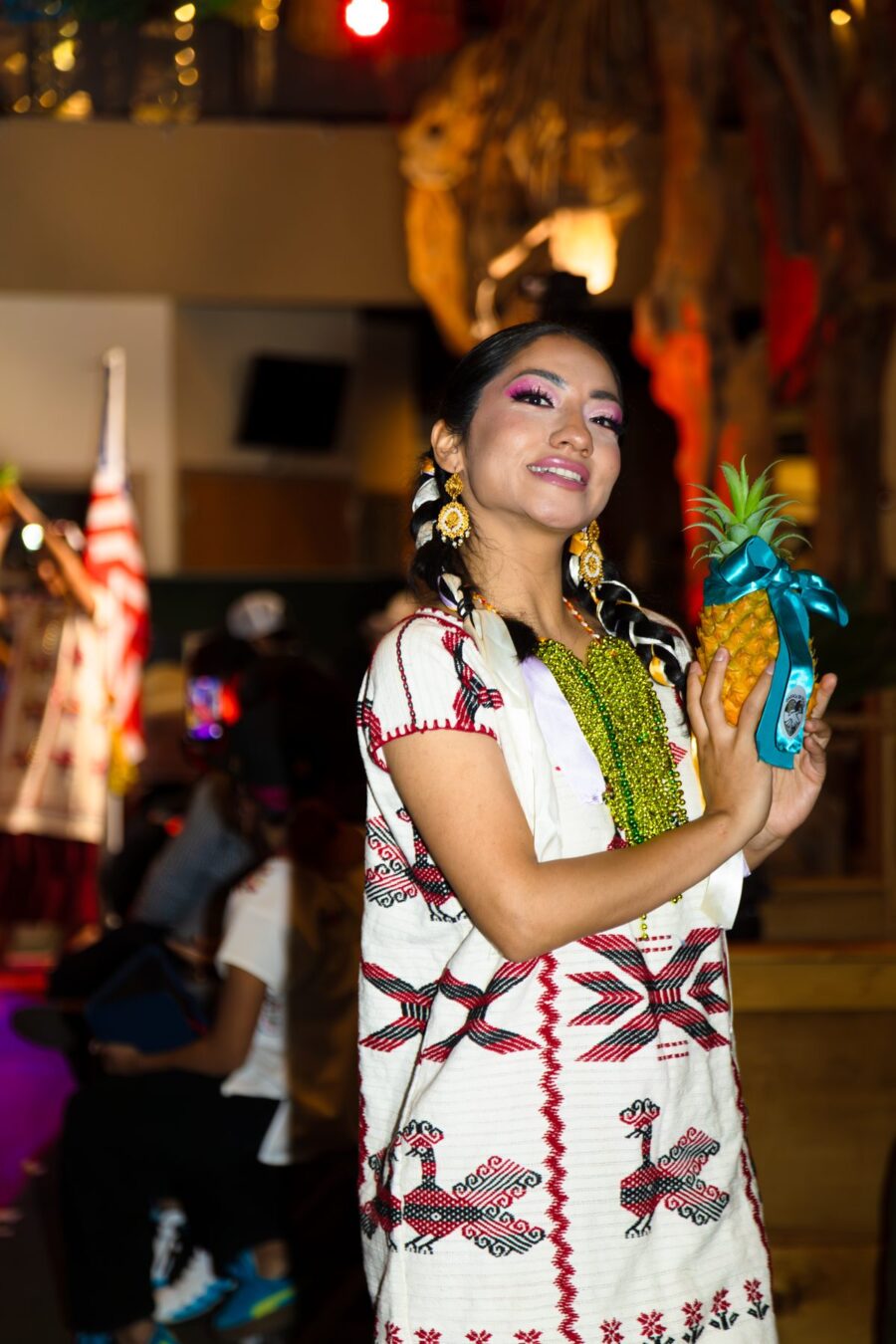 Una modelo con una prenda blanca tejida de diseños rojos y negros sostiene una piña decorada con un listón
