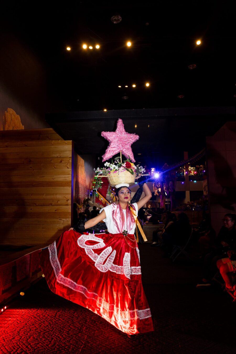 Una modelo con falda roja equilibra una canasta con flores y una estrella rosa mientras posa en la pasarela
