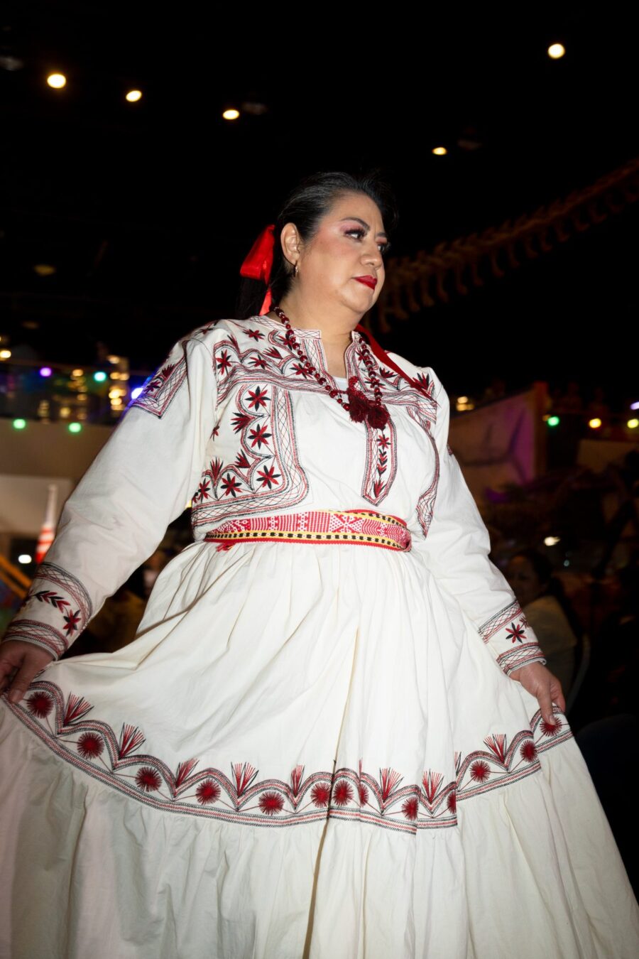 Una modelo presenta un vestido blanco con bordados rojos y negros bajo las luces de la pasarela