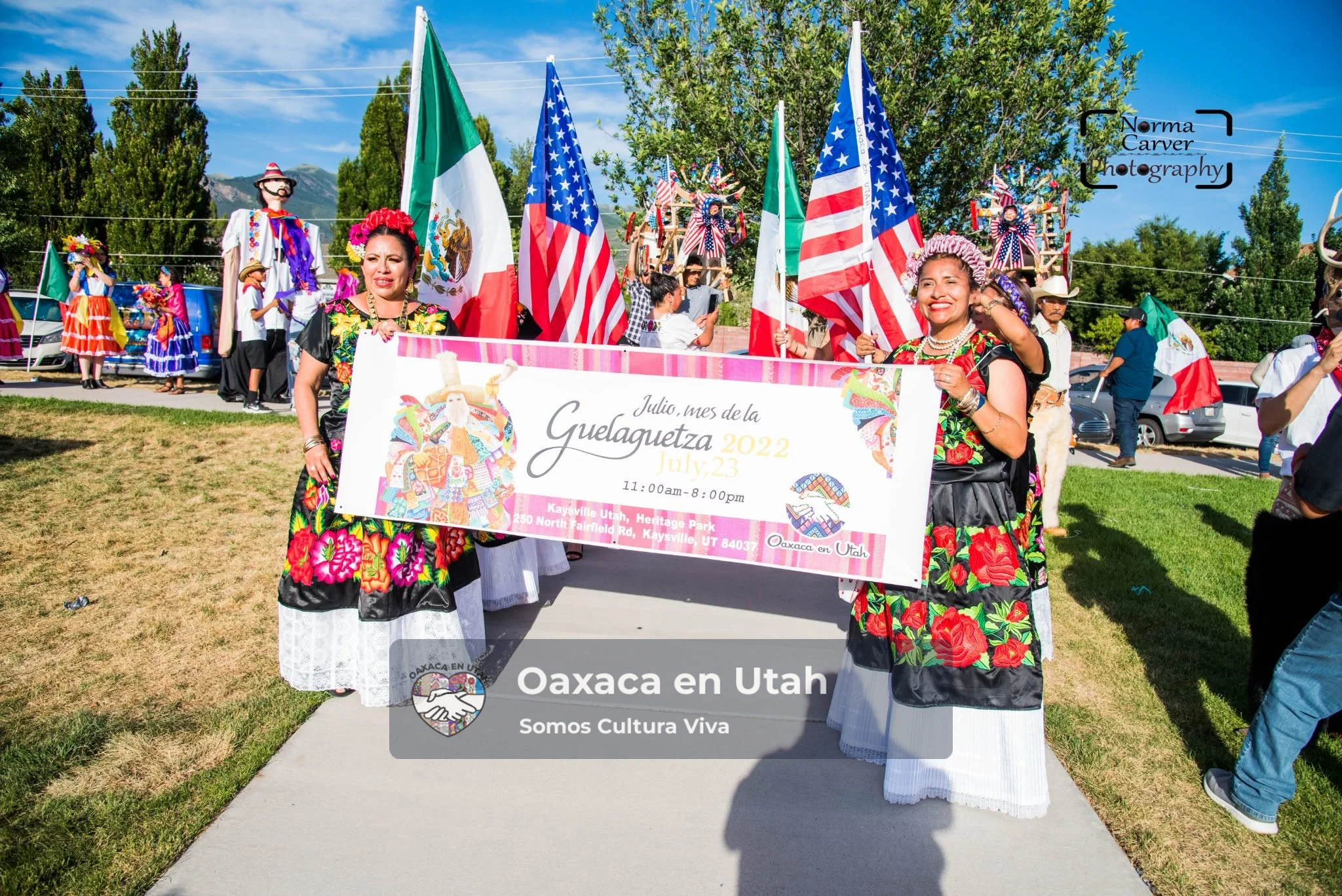 Dos mujeres llevan el cartel de La Guelaguetza 2022 entre banderas de México y Estados Unidos en Heritage Park