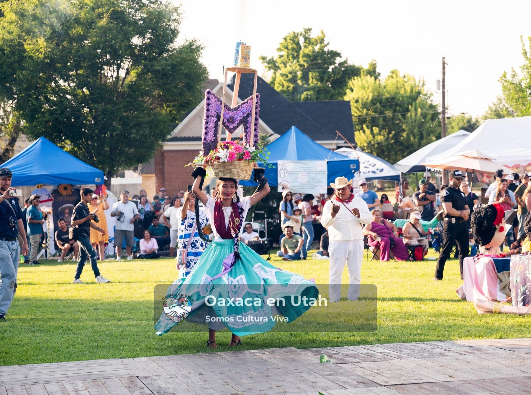 A participant in a turquoise skirt and tall decorated headpiece moves across an outdoor festival lawn