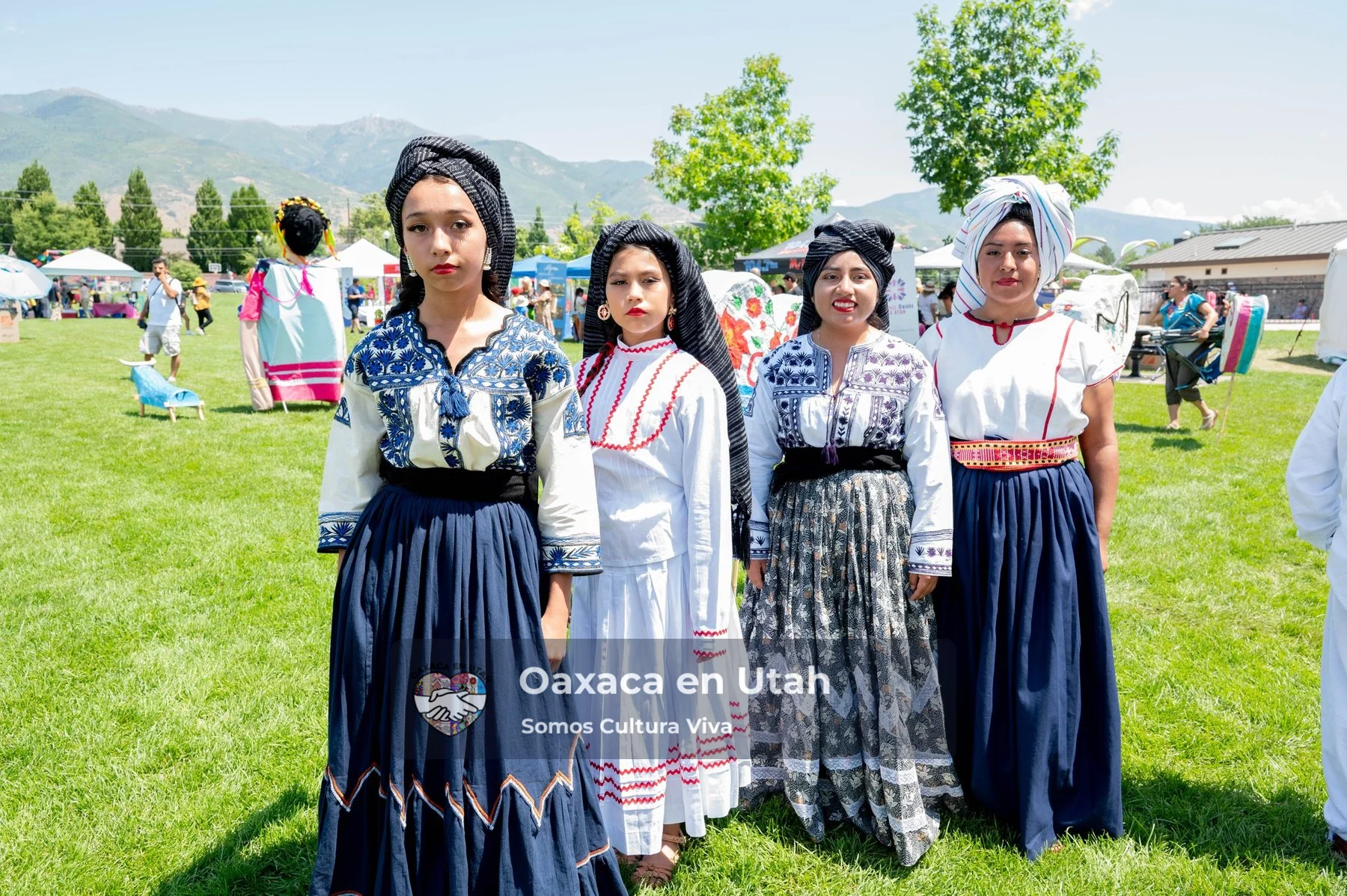 Four participants in black, white, and blue dresses stand on a lawn with mountains behind them