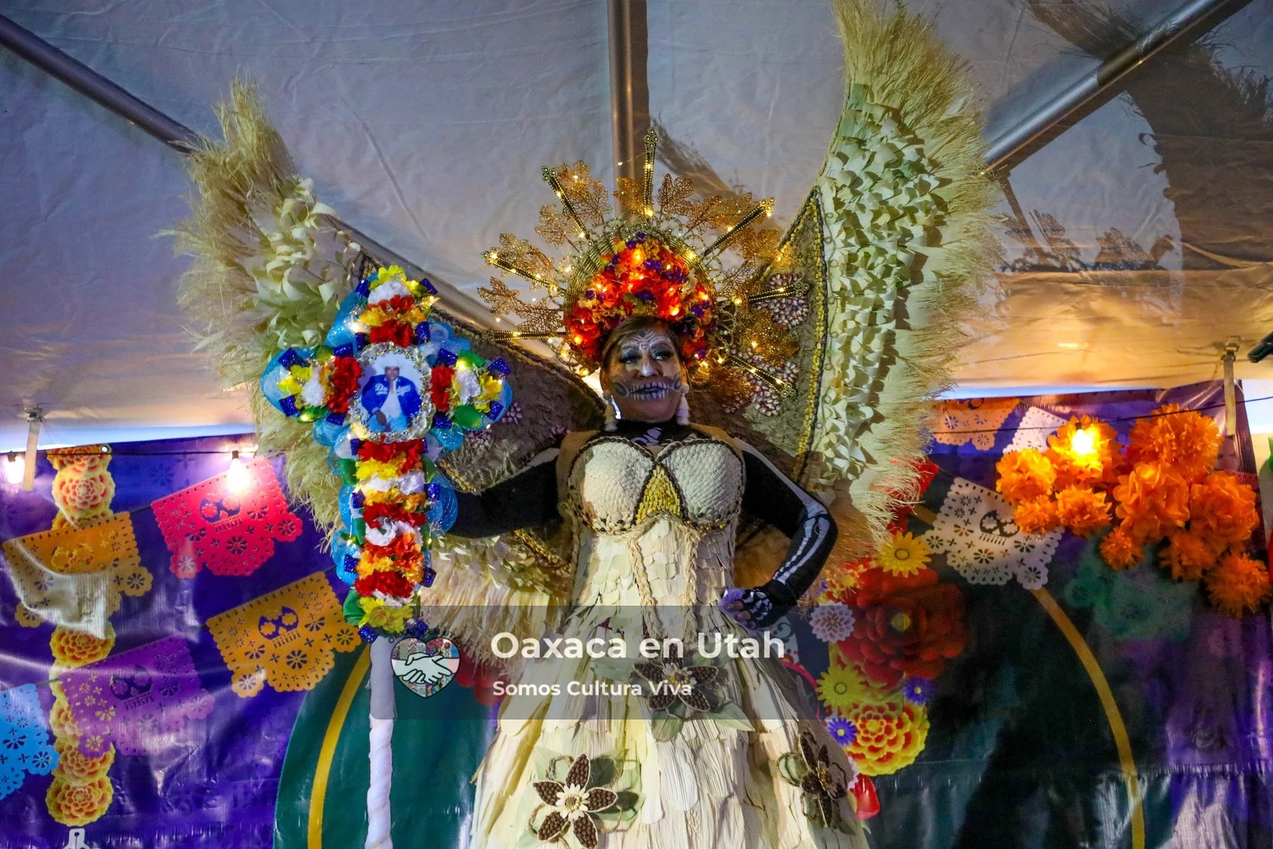 A performer in a broad feathered backpiece stands before a colorful altar display