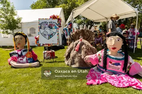 Two large colorful figures sit on the grass beside an Oaxaca en Utah banner