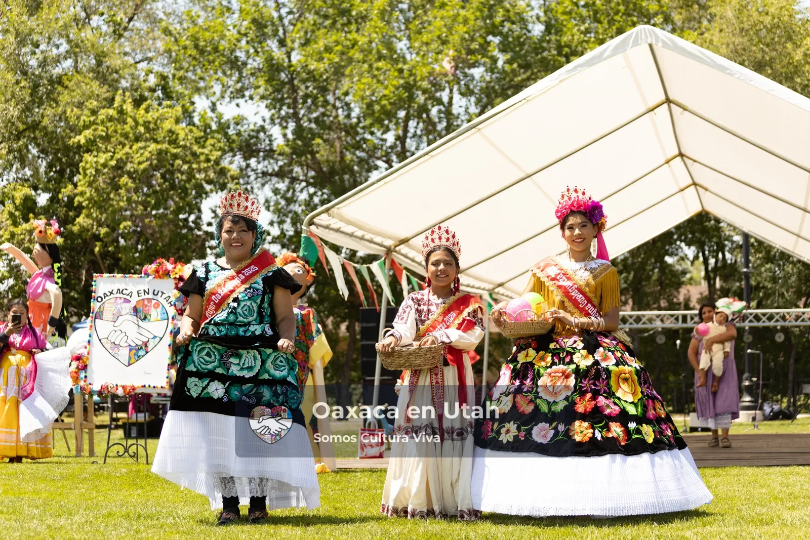 Tres participantes con vestidos bordados están juntas junto al escenario